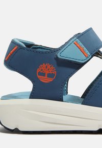 Sininen sandaali säädettävillä remmeillä, vaahtomuovipohjalla ja kumipohjalla; siinä on oranssi puu-logo ja erottuvat vaaleansiniset yksityiskohdat.