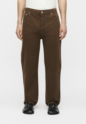 Καφέ παντελόνι με ίσια γραμμή που φοράει ένα όρθιο άτομο με μαύρα υφαντά παπούτσια χωρίς κορδόνια και λευκό τοπ, κομμένο στη μέση.