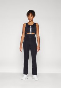 Black sports bra with "NEBBIA" logo and cut-out detail, paired with high-waisted black leggings. Smooth, stretchy fabric. White athletic shoes.