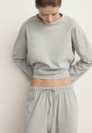 Woman in light grey long-sleeve cropped top and matching drawstring pants, looking down with hands behind her back.