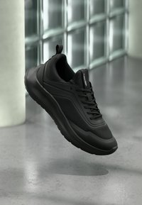 Μαύρο sneaker Calvin Klein με διχτυωτά και στερεά πάνελ, κορδόνια και θηλιά έλξης, αιωρούμενο πάνω από γυαλισμένο τσιμεντένιο πάτωμα κοντά σε γυάλινους τοίχους.