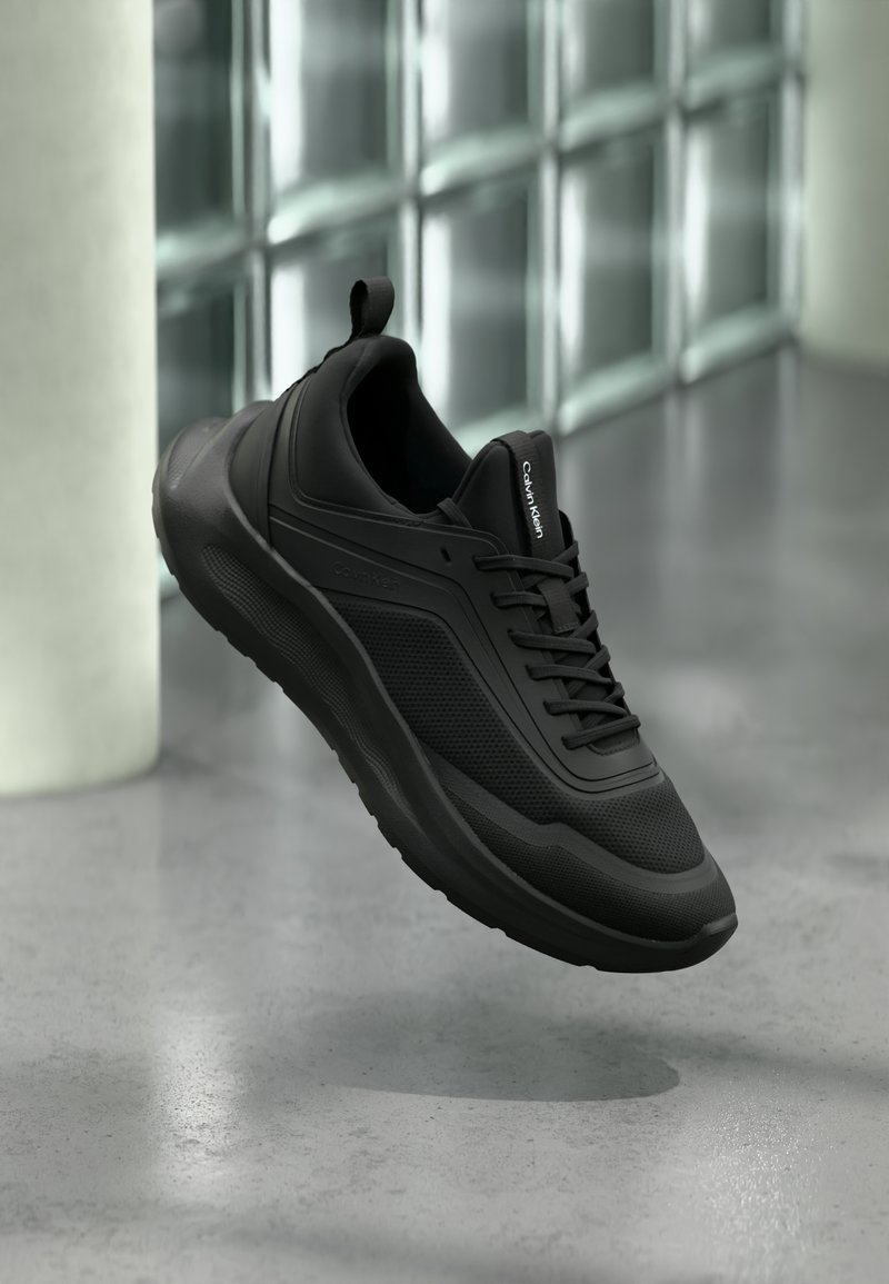 Μαύρο sneaker Calvin Klein με διχτυωτά και στερεά πάνελ, κορδόνια και θηλιά έλξης, αιωρούμενο πάνω από γυαλισμένο τσιμεντένιο πάτωμα κοντά σε γυάλι νους τοίχους.