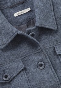 Chaqueta gris de felpa con cuello, dos bolsillos en el pecho y cierres de botón. Presenta un forro interior suave y botones de plástico negro.