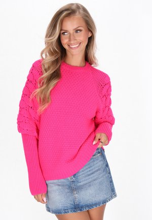 Roze gebreide trui met een textuurlichaam, geribde mouwen en open latticepatronen op de schouders. Gecombineerd met een blauwe denim rok.