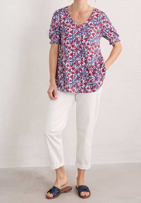 Top con estampado floral en rojo, azul y blanco con escote en V, mangas cortas abullonadas y bolsillos laterales, combinado con pantalones blancos de puño enrollado y sandalias marinas.