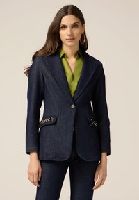 Blazer in denim blu scuro con vestibilità su misura, rever a scollatura, chiusura con bottoni dorati e dettagli decorativi sulle tasche. Abbinato a una camicia verde.