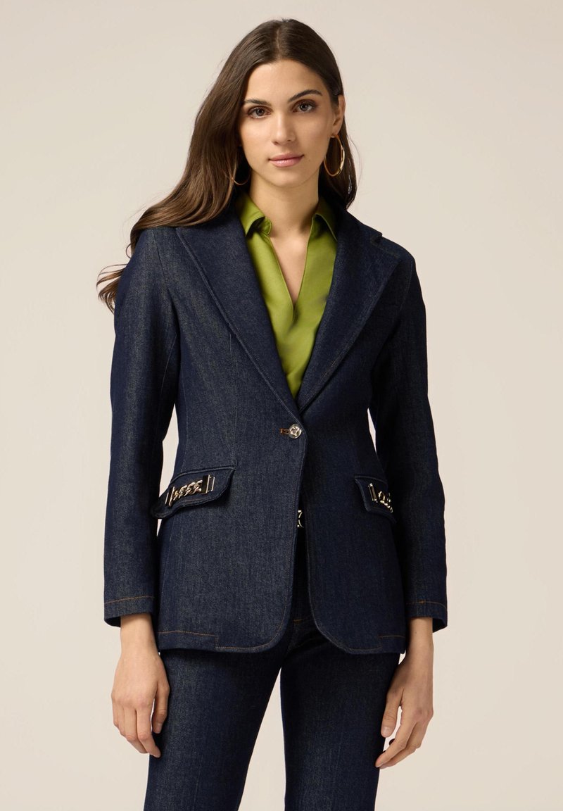 Blazer in denim blu scuro con vestibilità su misura, rever a scollatura, chiusura con bottoni dorati e dettagli decorativi sulle tasche. Abbinato a una camicia verde.