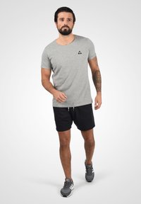 Camiseta de algodón gris con cuello redondo, parche de logo negro, combinada con pantalones cortos negros y zapatillas deportivas grises. El modelo está caminando.