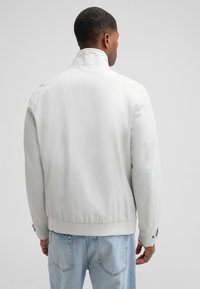 Weiße Bomberjacke mit glatter Oberfläche, Reißverschluss und geripptem Saum. Mit Schulterakzenten und Druckknopfmanschetten. Kombiniert mit hellblauen Jeans.