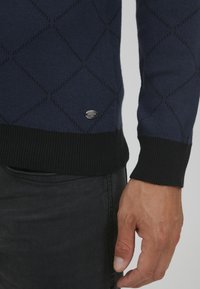 Navy knit sweater with diamond pattern, featuring black ribbed cuffs and hem. A small silver logo tag is visible on the front left side.