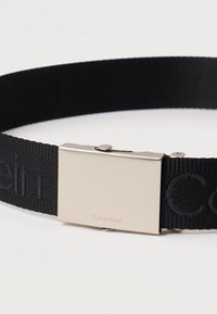 Μαύρη υφασμάτινη ζώνη με ματ ασημένιο ορθογώνιο κούμπωμα, με χαραγμένο το λογότυπο "Calvin Klein", δεμένη σε μια απλή ανοιχτόχρωμη επιφάνεια.