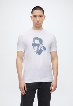 Νέος άνδρας με κοντά μαλλιά που φοράει λευκό μπλουζάκι με μια στυλιζαρισμένη μπλε εικόνα ενός άνδρα με γυαλιά ηλίου και αλογοουρά, συνδυασμένο με σκουρόχρωμα τζιν.