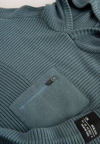 Gros plan d'un pull en maille bleu texturé avec une poche poitrine zippée et une étiquette noire avec texte blanc sur la manche.