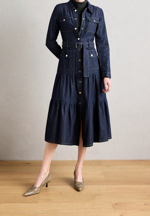 Femme portant une robe midi en denim bleu foncé ceinturée, avec une patte de boutonnage à l’avant, des poches poitrine et hanches, associée à des talons bruns à bout pointu.