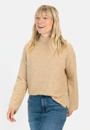 Beige gebreide trui met een hoge hals en lange mouwen, met een relaxte pasvorm en subtiele textuur, gecombineerd met blauwe spijkerbroek.