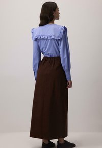 Femeie cu spatele îmbrăcată cu o bluză albastră cu guler volan și o fustă lungă maro, stând în fața unui fundal simplu de culoare deschisă.