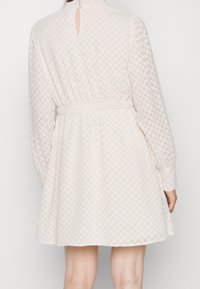 Vestido beige claro con mangas largas, cinturilla elástica, tela texturizada con un sutil patrón geométrico y detalle de escote en forma de ojo de cerradura en la parte posterior.