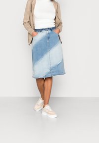 Denimkjol med diagonala ljusa och mörka blå paneler, medellång med en lätt A-linjeform, bärs med beige sneakers och en vit topp.