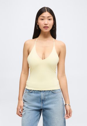 Femme aux longs cheveux foncés, portant de grandes boucles d'oreilles créoles, un haut dos nu jaune pâle et un jean bleu clair, debout devant un fond uni.