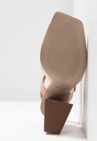 Podpatky a podrážka sandálů s texturovanými úchyty, béžová barva, označeno "Vyrobeno ve Španělsku", opřeno o bílé zdi a povrch.