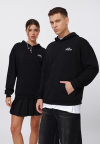 Sudaderas negras con bolsillos delanteros, que presentan el logo de "Los Ángeles". Modelo con una falda negra con pliegues y jeans oscuros, ambos en una paleta monocromática.
