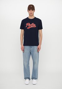 Jūras flotes kokvilnas T-krekls ar sarkano "Polo" logo, savienots ar gaiši zilo džinsām un baltām sporta apaviem. Brūna cepure papildina tērpu.