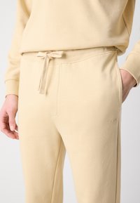 Pantaloni jogger din bumbac bej, cu talie ajustabilă prin cordon, buzunare laterale și un logo discret embosat pe piciorul inferior. Textură netedă.