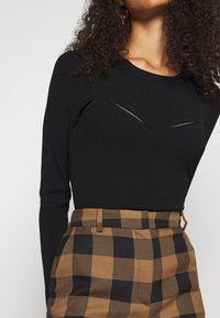 Schwarzes, langärmliges Rippstrickoberteil mit einem Chevron-Ausschnitt, kombiniert mit braunen und schwarzen, karierten, hoch taillierten Hosen.