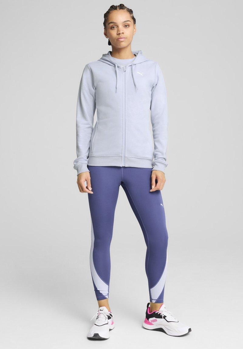 Helle graue Zip-Hoodie mit Kordelzügen, kombiniert mit marineblauen Leggings mit weißen Akzenten. Das Modell trägt weiße und rosa Sportschuhe.