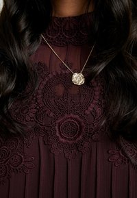 Femme portant une robe en dentelle bordeaux foncé avec des motifs floraux et un collier médaillon en or sur une fine chaîne.