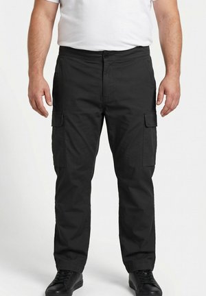 Man draagt zwarte cargobroek met zijzakken, wit t-shirt en zwarte sneakers, staand tegen een effen lichte achtergrond.