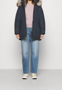 Granatowa parka z futrzanym obszyciem kaptura, zapinana na zamek i zatrzaski, noszona na jasnoróżowym swetrze, połączona z jasnoniebieskimi spodniami o prostych nogawkach i wzorzystymi sneakersami.