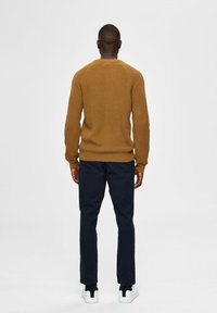 Hnědý pletený svetr s texturovaným vzorem, doplněný tmavě modrými kalhotami a bílými teniskami, z pohledu zezadu.