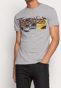Man wearing light gray Pepe Jeans London graphic t-shirt and black pants, hand in pocket against white background.