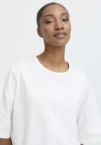 Kvinde med mørk hud og kort hår iført en enkel hvid oversized t-shirt, der ser selvsikkert ind i kameraet mod en ensfarvet baggrund.