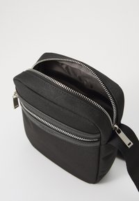 Sac bandoulière noir à fermeture éclair, fabriqué en tissu texturé. Il dispose d'une poche avant avec une fermeture éclair en argent poli et une sangle réglable.