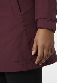 Chaqueta impermeable burdeos con tejido texturizado, que presenta un puño doblado y el logo "HELLE TECH®" en el dobladillo. Proporciona protección contra los elementos.