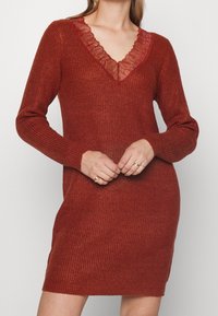 Robe en maille côtelée de couleur rouge profond avec des manches longues et un décolleté en V bordé de dentelle. Coupe ajustée avec un ourlet droit, dotée de fentes sur les côtés.