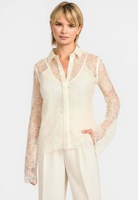 Transparent blondebluse i lys creme med lange, flagrende ærmer, krave og knaplukning, prydet med blomsterdesign.