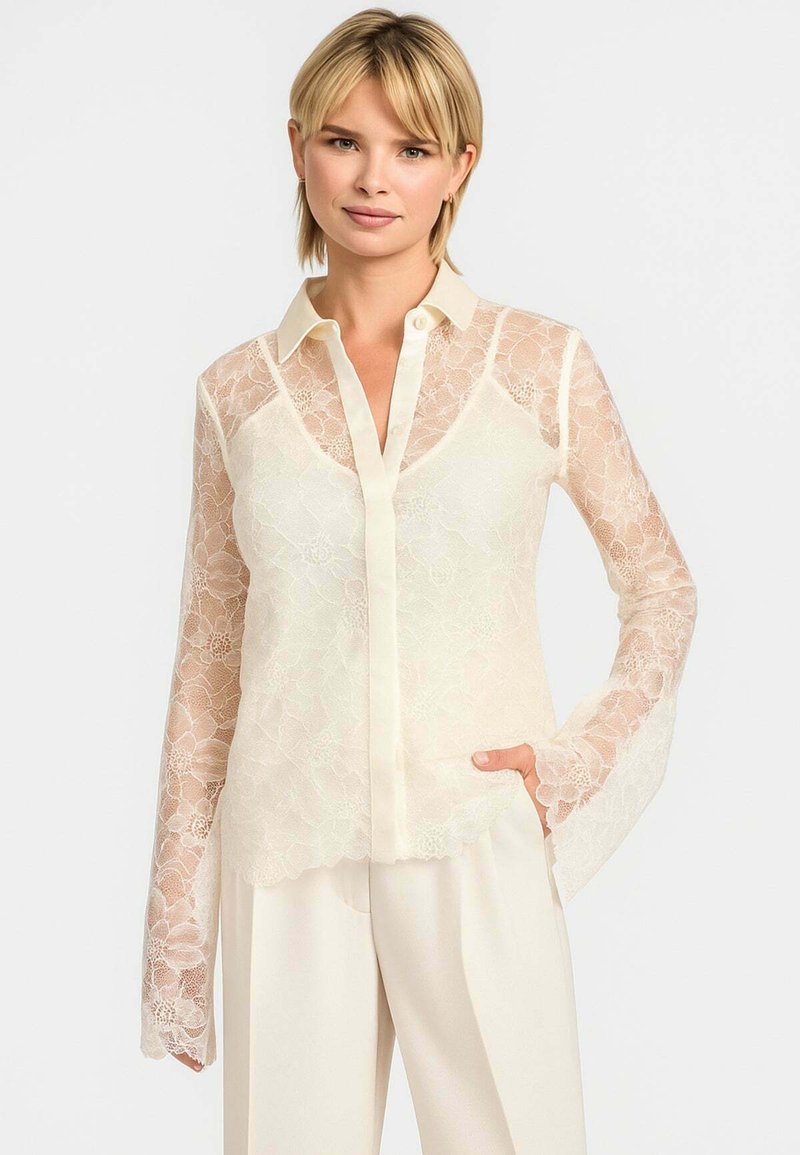 Transparent blondebluse i lys creme med lange, flagrende ærmer, krave og knaplukning, prydet med blomsterdesign.