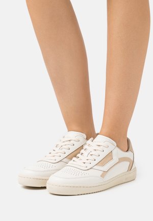 Ben iført hvide og beige lave lædersneakers med snørebånd, perforeret tå og gummisål på en ensfarvet lys baggrund.
