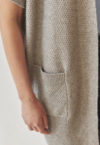 Cardigan en tricot beige à manches courtes, présentant une surface texturée et une poche avant visible. Ourlet côtelé aux manches et en bas.