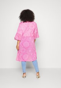 En person med lockigt hår bär en rosa mönstrad kimono över ljusblå jeans och vita klacksandaler, vänd bort mot en neutral bakgrund.