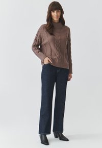 Suéter marrón grueso de punto con un alto cuello y patrones de trenza, combinado con pantalones vaqueros de pierna ancha en azul oscuro y botines negros.