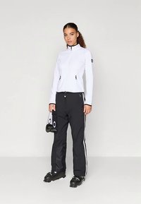 Kobieta ubrana w białą kurtkę zapinaną na zamek i czarne spodnie narciarskie, trzymająca czarne gogle narciarskie, stojąca na jednolitym jasnoszarym tle.