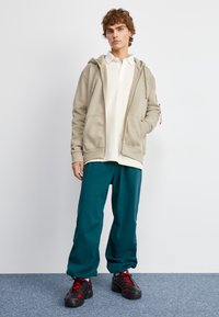 Bežna zip-up sweatshirt, bijela polo majica, tirkizne hlače i crne cipele s crvenim detaljima. Ležeran kroj, opušten fit, s prednjim džepovima i vezicom.