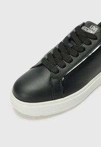 Zwart leren sneaker met ronde neus, textuur zwarte veters, witte rubberen zool en zilveren accenten aan de zijkant. Logo gedrukt op de tong.