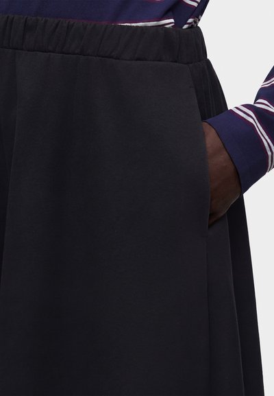 Άτομο που φοράει μαύρη φούστα με ελαστική μέση με το δεξί χέρι στην πλαϊνή τσέπη, σε συνδυασμό με μπλε σκούρο και λευκό ριγέ μακρυμάνικη μπλούζα.