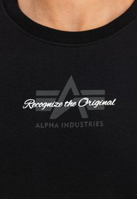 Melnā jakā ar ribotu apkakli, kurā ir balti uzraksti "Atpazīsti oriģinālu" un "ALPHA INDUSTRIES" izteiksmīgā fontā.