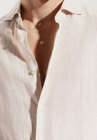 Zbliżenie na osobę w białej, lekko przezroczystej koszuli z guzikami rozpiętymi na górze, odsłaniającej część szyi i klatki piersiowej.